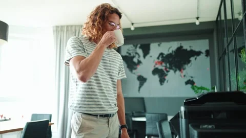 Young long haired man drinking coffee and prints on printer in office 動画素材 244258259