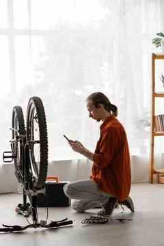 Young long-haired man in orange shirt repairing bicycle chatting online wit.. Stock Photos