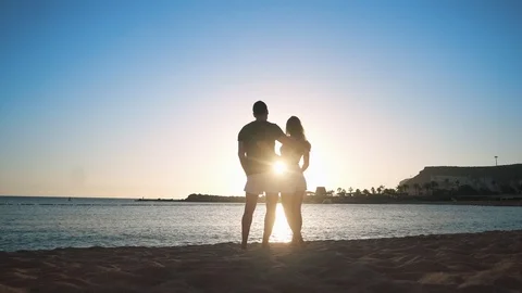 A young loving couple is standing on a sandy beach by the sea at sunset, holding Stock Footage 104203177