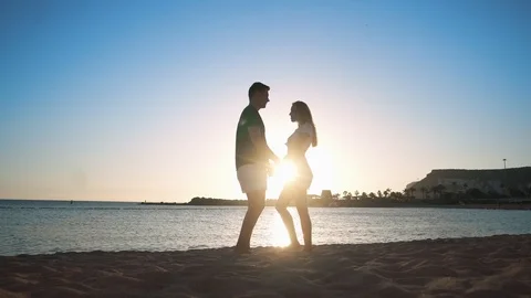 A young loving couple is standing on a sandy beach by the sea at sunset, hugging Stock Footage 104203536
