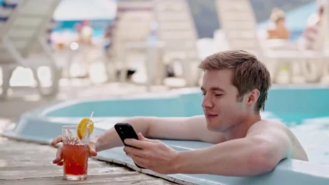 Young macho man browsing social media in swimming pool and drinking aperol Stock Footage 256802126