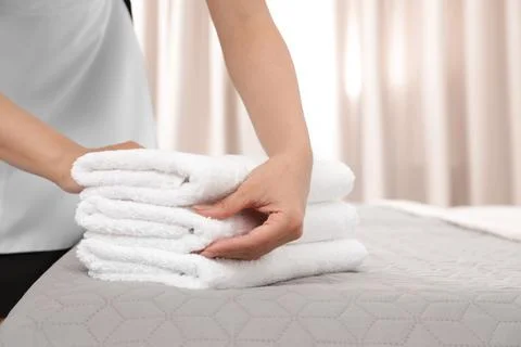 Young maid putting stack of fresh towels on bed in hotel room, closeup 스톡 사진