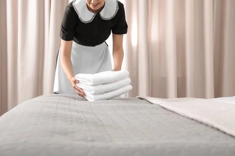 Young maid putting stack of fresh towels on bed in hotel room, closeup 스톡 사진