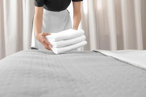 Young maid putting stack of fresh towels on bed in hotel room, closeup 스톡 사진