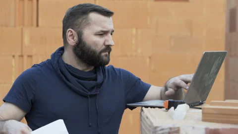 Young male constructor comparing data on laptop and blueprints at construction Stock Footage 87455912