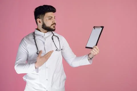 Young, male dctor, intern standing, looking aside, holding folder, raising hand Stock Photos