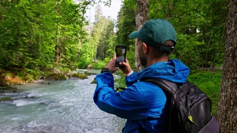 Young male explorer with a backpack filming a wild mountain stream in a lush Stock Footage 340997351