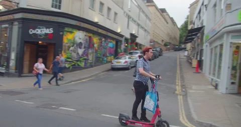 Young Male Hipster Holding Bag Riding Electric Scooter Up Park Street in Bristol Stock Footage