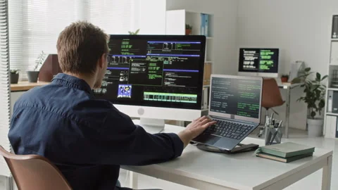 Young Male IT Specialist Debugging Code on Laptop and Desktop Computer in Office Stock Footage 330124647