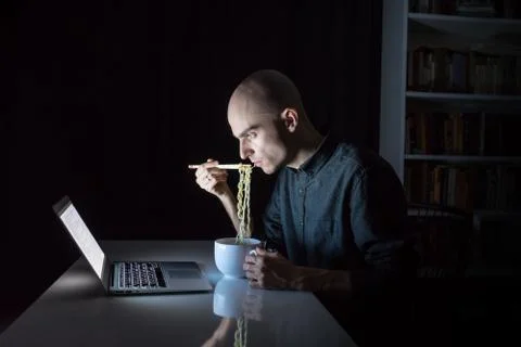 Young male at laptop computer eats instant ramen noodles with chopsticks Stock Photos
