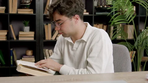 Young male reader in white sweater examines pages of an open book while seated Stock Footage 330805098