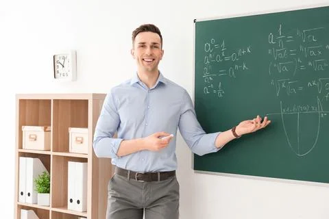 Young male teacher explaining math formulas written on blackboard in classroo Stock Photos