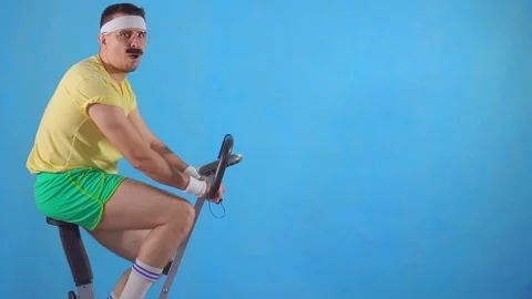 Young man from 80's with mustache on exercise bikeon blue background showing Stock Footage 107798616