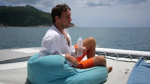 A young man admires the view, sitting on a yacht and drinks clean water from a Stock Footage 94889627