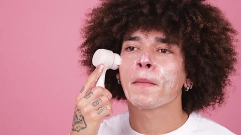 A young man with an afro hairstyle using a circular facial cleansing brush on 스톡 동영상 269363065