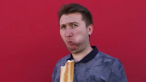 A young man against background of a red wall chews and bites off bread Stock Footage 137532768