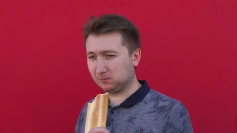 A young man against background of a red wall chews and bites off bread Stock Footage 137532792