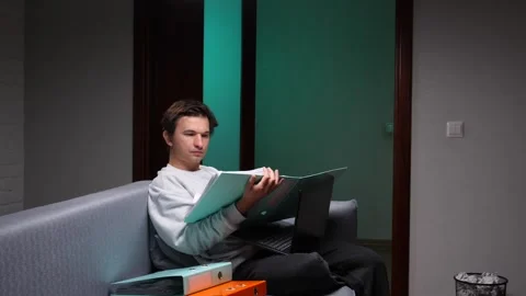 Young man analyzing papers and working on a laptop while sitting comfortably on Stock Footage 347241372