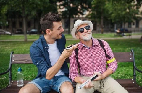Young man and blind senior sitting on bench in park in city, using smartphone. Stock Photos