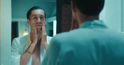 Young man applying moisturizer cream on face skin looking in bathroom mirror Stock Footage 244069401