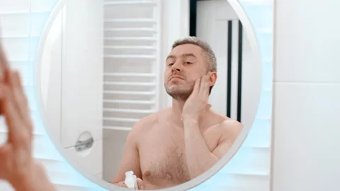 Young man applying moisturizer cream on face skin looking in bathroom mirror Stock-Footage 254069499