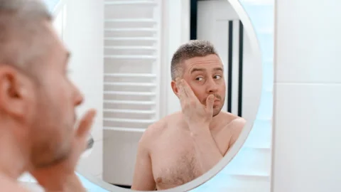 Young man applying moisturizer cream on face skin looking in bathroom mirror Video stock 259635202