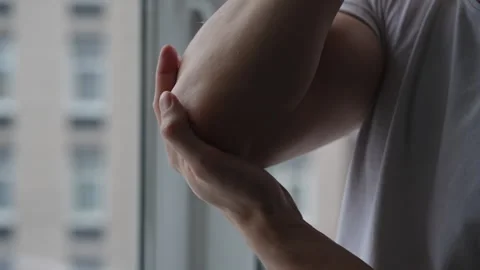Young man applying moisturizer cream on elbow standing by window at home Stock Footage 268392304