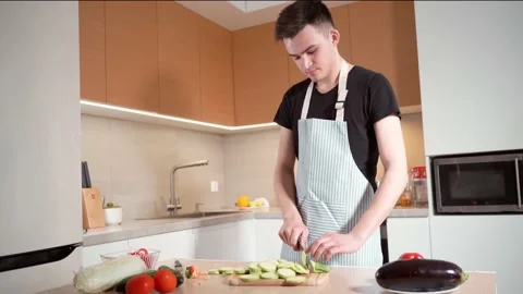 A young man with an apron in the kitchen cuts a zucchini on a cutting board. Stock Footage 153456224