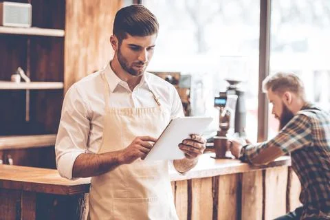 Young man in apron using his digital tablet while standing at cafe 库存照片