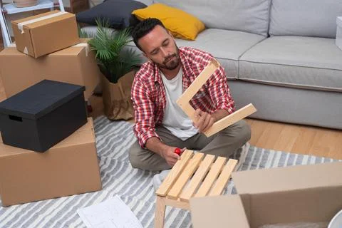 Young man assembles DIY shelf amidst moving carton boxes, adding warmth to home Stock Photos