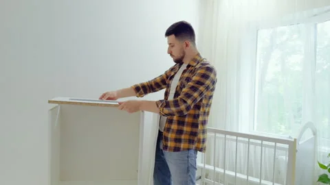 Young man assembles white wooden furniture for the child's room. Young father Stock Footage 282400696