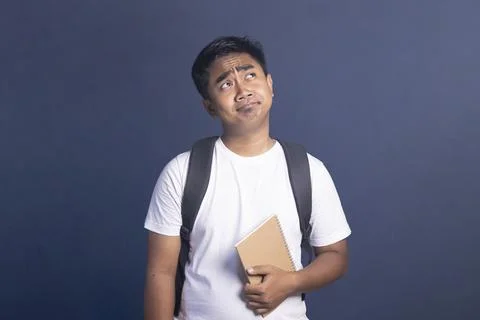 Young man with a backpack holding a notebook looking upwards with a worried.. Stock Photos