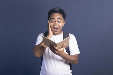 Young man with a backpack holding an open notebook while touching his face .. Stock Photos