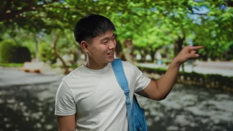 Young man with a backpack pointing and smiling outdoors in a lush green par.. Video stock 306837631
