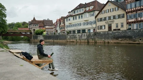 Young man with backpack sits dangling legs on a wooden pier near water and uses 스톡 동영상 115948255