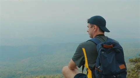 Young man with backpack sitting on mountain top, enjoying serene view Stock Footage 281011154