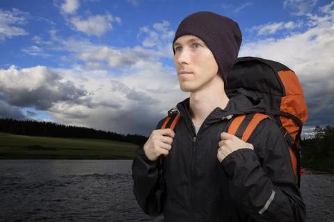 Young man with a backpack stands in front of a river. Stock Photos