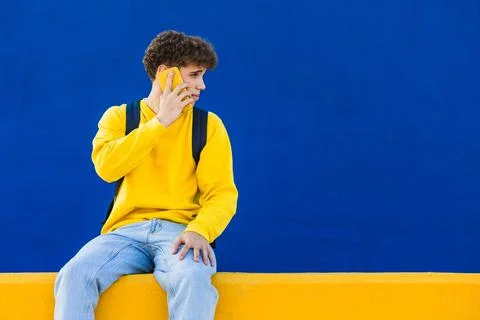 Young man with backpack using smartphone sitting on yellow wall with blue Stock Photos