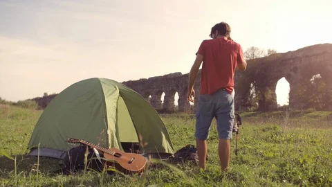 Young man backpacker outside camping tent picks up guitar and walks on grass in Stock Footage 86276869