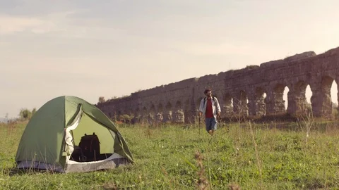 Young man backpacker outside camping tent walks barefeet with sticks on grass in 스톡 동영상 86283384