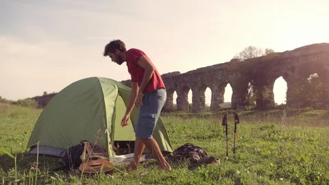 Young man backpacker outside camping tent picks up guitar and walks on grass in 스톡 동영상 121337234