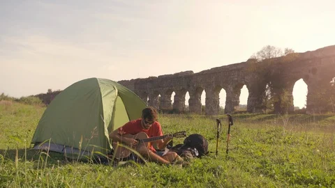 Young man backpacker playing guitar outside camping tent sitting on grass in Stock-Footage 86268661