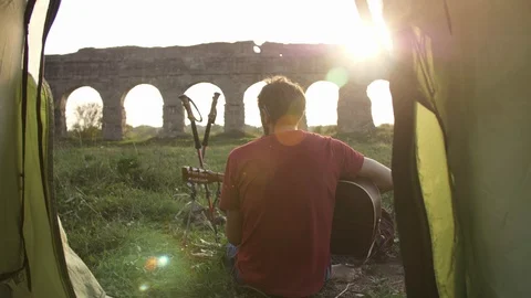 Young man backpacker playing guitar outside camping tent sitting on grass in 스톡 동영상 115192765