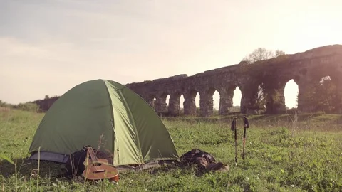 Young man backpacker wakes up exits camping tent in front of roman aqueduct Video stock 121337559