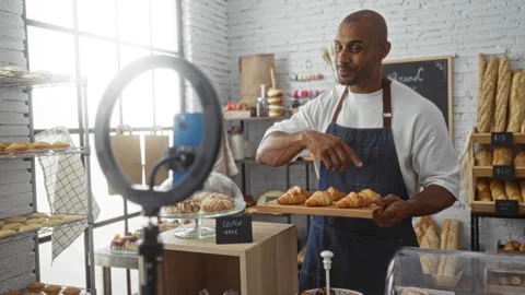 Young man in bakery holding tray of croissants while filming tutorial with .. 動画素材 289308673