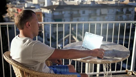 A young man on the balcony drinks a drink and works on his laptop Stock Footage 122853022