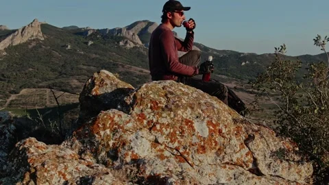 A young man in a baseball cap is resting on the top of a mountain and drinking f Stock-Footage 150592683