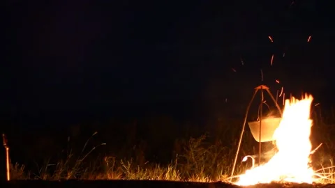 Young man basks by the fire preparing food in a pot on a tripod Stock Footage 131956285