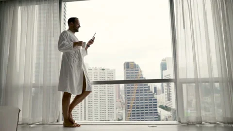 Young man in bathrobe browsing tablet standing by window with city view, 4K Stock Footage 127972718