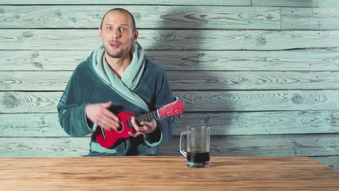Young man in a Bathrobe is drinking beer, singing and playing ukulele for his 스톡 동영상 104465900